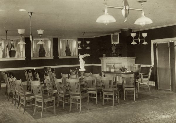 Interior view of the Neenah Public Library. The library opened in 1904 and was funded with a combined donation of $20,000 from the citizens of Neenah and Andrew Carnegie. Reverse of the cardboard backing reads: "Womans' Club Room." A group of chairs face a table near a fireplace at one end of the room. Near the far wall is a statue of Winged Victory, and along the wall are three small windows with curtains.