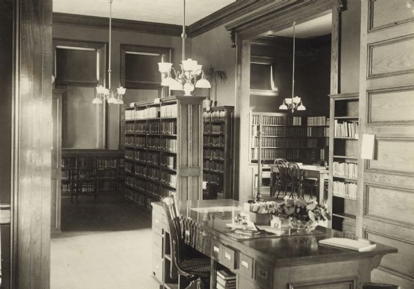 Interior view of the Neenah Public Library. The library opened in 1904 and was funded with a combined donation of $20,000 from the citizens of Neenah and Andrew Carnegie. Reverse of the cardboard backing reads: "A glimpse of stack room and reference room." In the foreground is a librarian's desk with card catalog drawers facing the librarian. Beyond the desk are bookshelves, a reading table, and chandeliers.