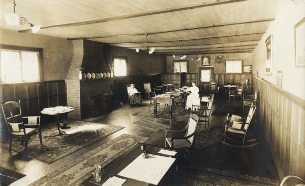 Interior view of the Oconomowoc Public Library. The library was funded by a combined gift of $4,500 from Mrs. Geo Bullen and Mr. P. D. Armour, Sr. Photograph is stamped in lower left: "Manger Oconomowoc, Wis." There is a fireplace with a sign above it that reads: "Please be quite." In the foreground is a librarian's desk, and there are reading tables and rocking chairs throughout the long, narrow room. On the right is a fireplace. Two women are reading. A sign on the mantel says: "Please Be Quiet."
