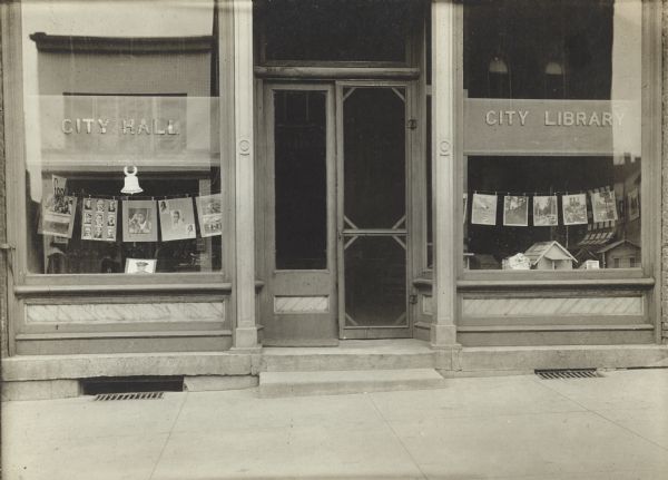 Exterior view of show windows of the Prairie du Chien City Library. The cardboard backing reads: "Prairie du Chien, 1918." Reverse of the cardboard mounting reads: "Prairie du Chien Library Show Windows." Stamped on the cardboard mounting are the word: "Nichols." The storefront library shared space with city hall. Photographs are clipped to strings hanging across the windows.
