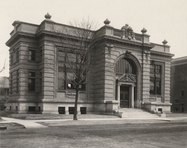 Exterior view of the Racine Public Library. The building was funded with a donation from Andrew Carnegie. Reverse of the cardboard backing reads: "1904, cost $50,000 Mauran, Russel, Garden, St Louis, architect." Above the main entrance it reads: "Free To the People." Prominent elements of the photo: facade, brick building, large arch over the main entrance, columns, steps, sidewalk.