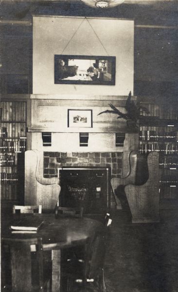 Interior view of Racine Public Library Branch. In the room are two wooden benches on either side of a fireplace. A reading table is in the foreground, and on the back wall are bookshelves.
