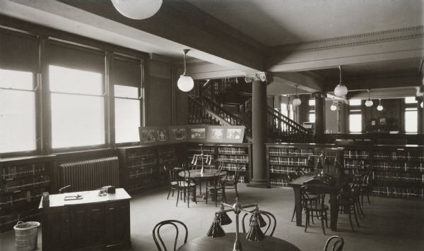Interior view of the Racine Public Library. Reverse of cardboard backing reads: "Part of Children's Room." In the foreground are reading tables with lamps, bookshelves, and a librarian's desk near three windows. In the background is a stairway to an upper floor.
