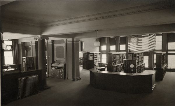 Interior view of the Racine Public Library. Reverse of cardboard backing reads: "Delivery Desk and Stacks." In the room is a large five-sided librarian's desk, and a card catalog. Behind the desk are bookshelves, and a U.S. flag hanging from the ceiling. There is a room on the left, separated by columns from the entryway.