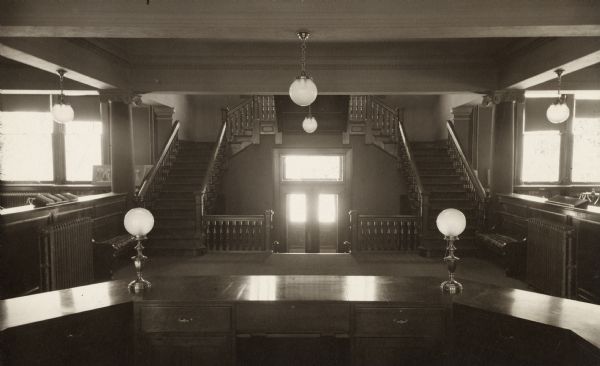 Interior view of the Racine Public Library. Reverse of cardboard backing reads: "Entrance from desk." In the foreground is a large five-sided librarian's desk, which faces a double wooden staircase to upper floor. Entrance doors are down one level. Two rooms can be seen on either side of the staircase.