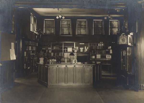 Interior view of the Rhinelander Public Library. The library opened in 1904 funded with a $15,000 donation by Andrew Carnegie. Several shelves are labeled, including: "New Books," "Fiction," "Seven Day Books, Cannot Be Renewed." There is a large librarian's desk, a card catalog, and bookshelves. Windows are above the bookshelves with the shades drawn. On the right is a grandfather clock.
