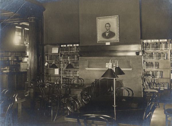 Interior view of the Rhinelander Public Library. The library opened in 1904 funded with a $15,000 donation by Andrew Carnegie. There is a portrait of Abraham Lincoln hanging over a fireplace, bookshelves are along a back wall. Reading tables and bentwood chairs are in the foreground. A second room with a large librarian's desk and card catalog is on the left beyond a carved column in an archway. 
