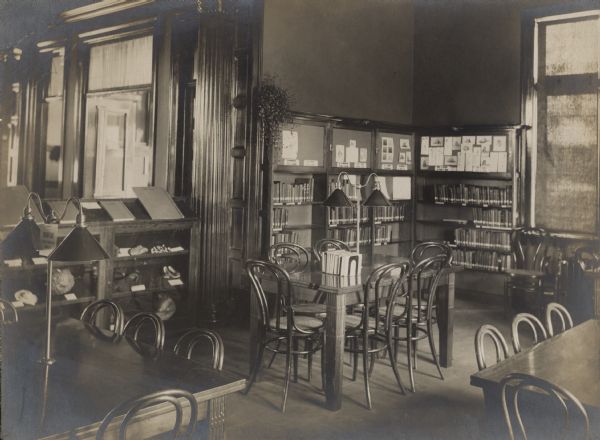 Interior view of the Rhinelander Public Library. The library opened in 1904 funded with a $15,000 donation by Andrew Carnegie. In the foreground are reading tables with reading lamps and bentwood chairs. Behind the tables are display cases and bookshelves.