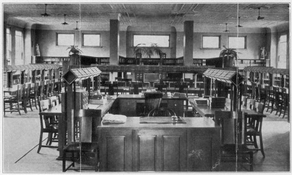 Interior view of the State Normal School Library in River Falls. Below the image it reads: "Beautiful New Library, State Normal School, River Falls, Wisconsin." In the large open room with windows and columns are two desks with tables on either side forming a central square area. In the background are long reading tables and chairs, bookshelves, and a card catalog.
