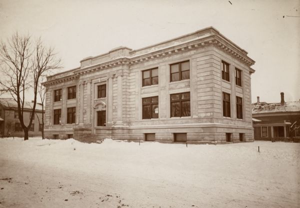 Exterior view of the Sheboygan Public Library in winter. The library opened in 1903. Reverse of the cardboard backing reads: "est 1897, Carnegie bldg, 1903. cost $35,000, Patton + Miller, Chicago, arch." The stone building has an arch over the entrance. The words "Pubic Library" are carved above a window.
