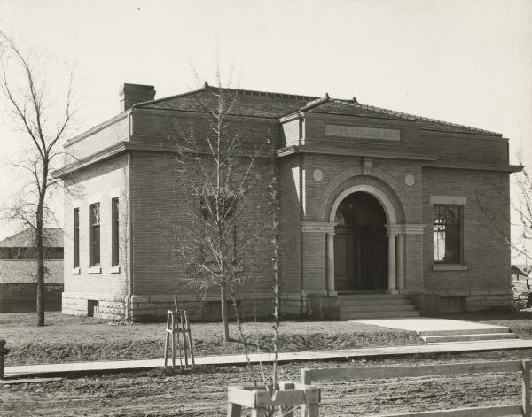Exterior view from road of the Stanley Public Library. The library opened in 1901 and was funded with a $15,000 donation from Mrs. S.F. Moon. Above the main entrance it reads: "Public Library." The hip roofed brick building has an arched porch over the entrance. New trees have protective wood supports, the grounds around the library is not yet landscaped, and the road is unpaved.