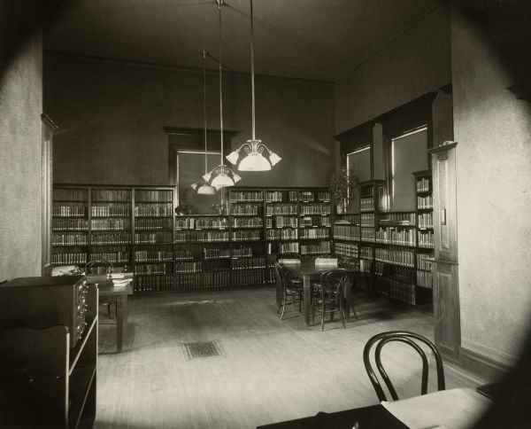 Interior view of the Stanley Public Library. The library opened in 1901 and was funded with a $15,000 donation from Mrs. S.F. Moon. In the room are reading tables with bentwood chairs, a small six-drawer card catalog, and bookshelves along the walls. Three lamps hang from the ceiling.