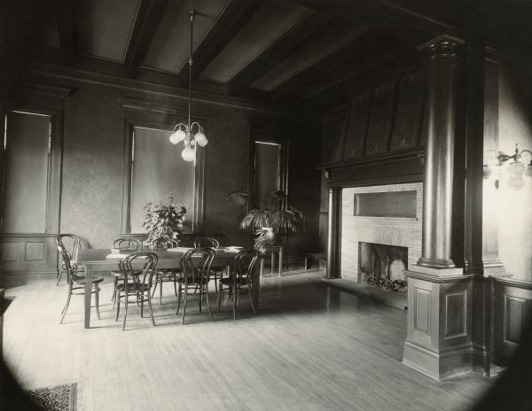 Interior view of the Stanley Public Library. The library opened in 1901 and was funded with a $15,000 donation from Mrs. S.F. Moon. In the room are a large reading table with bentwood chairs under a chandelier, potted plants, a sconce, and a fireplace with andirons. The tall ceiling has exposed beams, and a column is at the side of the entrance to the room. Over the fireplace is a plaque dedicating the library to Delos R. Moon.