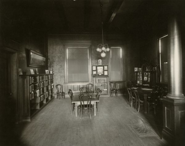 Interior view of the Stanley Public Library. The library opened in 1901 and was funded with a $15,000 donation from Mrs. S.F. Moon. In the room is a column on the right and a ceiling with exposed beams. There are reading tables with bentwood chairs, bookshelves along the walls, potted plants, a fern, a small children's reading table. Pictures of birds displayed on the bookshelves.
