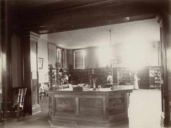 Interior view of the Stevens Point Public Library. The library opened in 1904 and was funded by a $22,000 donation from Andrew Carnegie. In the center, framed by a carved wood molding is a five-sided librarian's desk. On the left is a rocking chair. Behind the desk is a large room with windows that have venetian blinds. Three women and two boys are standing reading and browsing among the bookshelves which line the walls.