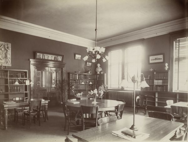 Interior view of the Stevens Point Public Library. The library opened in 1904 and was funded by a $22,000 donation from Andrew Carnegie. In the room are reading tables with lamps, bookshelves against a wall, and windows along the wall on the right. A man is seated at a table reading a book, and a boy stands reading a book on a book stand. On the far wall are closed double doors.