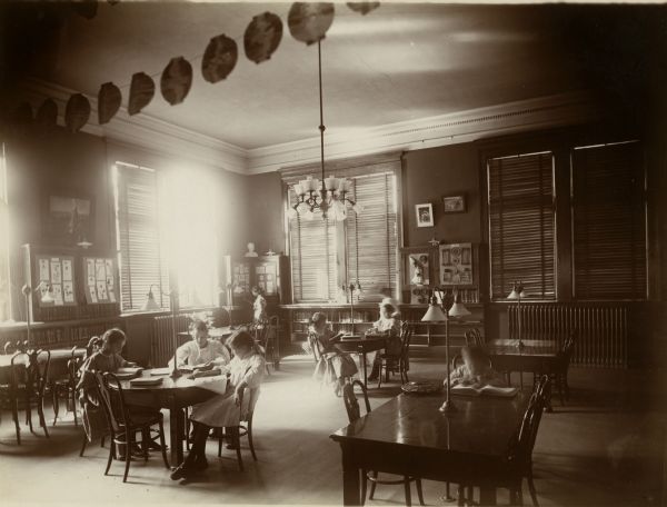 Interior view of the Stevens Point Public Library. The library opened in 1904 and was funded by a $22,000 donation from Andrew Carnegie. The children's Room has reading tables and chairs, a display case along one wall. Venetian blinds shade the tall windows. Children are reading at the tables. A string of faux Chinese lanterns is strung along the ceiling in the foreground. 
