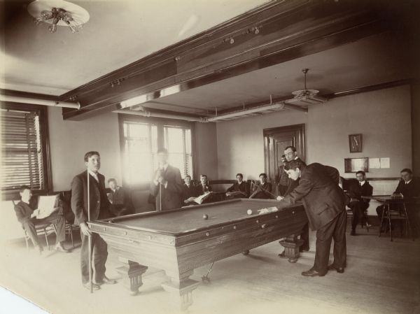 Interior view of the Stevens Point Public Library. The library opened in 1904 and was funded by a $22,000 donation from Andrew Carnegie. In the room is a pool table and young men are playing billiards. Other men sit along the wall reading newspapers.