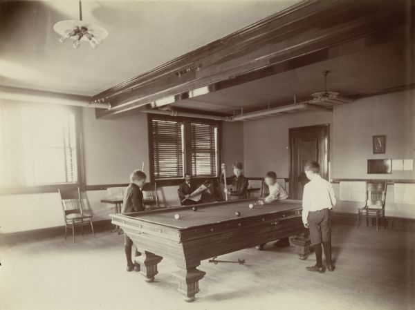 Interior view of the Stevens Point Public Library. The library opened in 1904 and was funded by a $22,000 donation from Andrew Carnegie. Young boys are playing billiards, while a man sits behind them reading a newspaper.