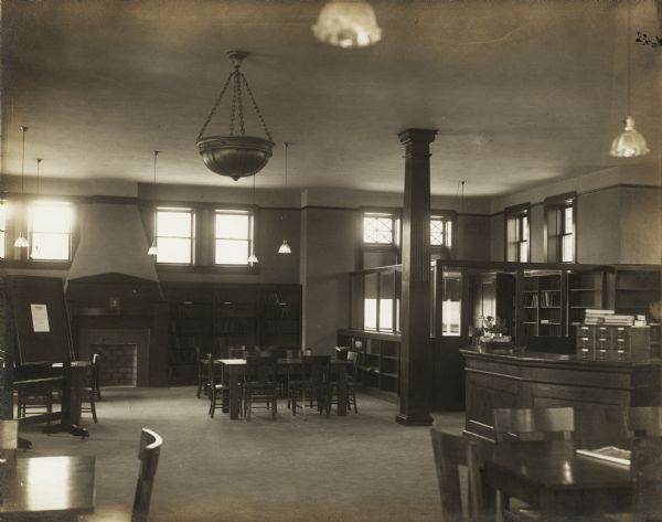 Interior view of the Sturgeon Bay Public Library. Library opened in 1913 with a gift of $12,500 from Andrew Carnegie. To the left of the photograph is a posting of: "The Rules." In the room is a librarian's desk with card tray, a nine-drawer card catalog, reading tables, bookshelves, and a fireplace. In the right corner beyond a carved wood column is an office separated by bookshelves and glass dividers.