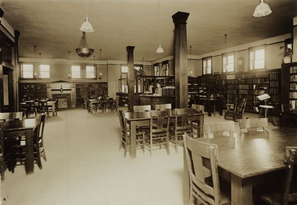 Interior view of the Sturgeon Bay Public Library. In the room are reading tables, two carved wood columns, a fireplace, bookshelves, and a nine-drawer card catalog. A woman (librarian?), stands at the desk. In the back corner is an office is an office separated by bookshelves and glass dividers.