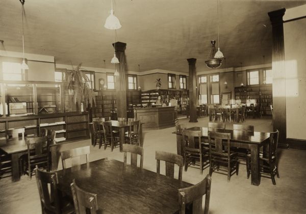 Interior view of the Sturgeon Bay Public Library. Library opened in 1913 with a gift of $12,500 from Andrew Carnegie. In the room are reading tables, bookshelves, and a card catalog. In the left corner of the room is an office separated by bookshelves and glass dividers. A  woman (librarian?) stands at the desk.