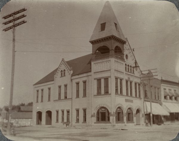 Exterior view from across road of the Sun Prairie Public Library. Building pictured is Sun Prairie City Hall. Located on the corner of Main Street and Bristol Street, the building was constructed in 1895. The library was located in the village council room until the Public Library, at 115 East Main Street, was constructed in 1924. There is a large square corner tower on the corner, with an arched balcony within the tower. Arched windows are on the first floor, and along the left side of the building are two large open garage doors.