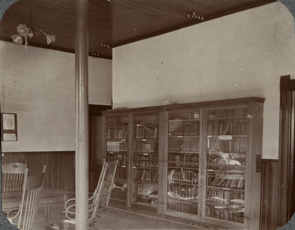 Interior view of the Sun Prairie Public Library. Room pictured is located within the Sun Prairie City Hall. Located on the corner of Main Street and Bristol Street, the building was constructed in 1895. The library was located in the village council room until the Public Library, at 115 East Main Street, was constructed in 1924. In the room are bookshelves with glass along a wall, and reading table with chairs.