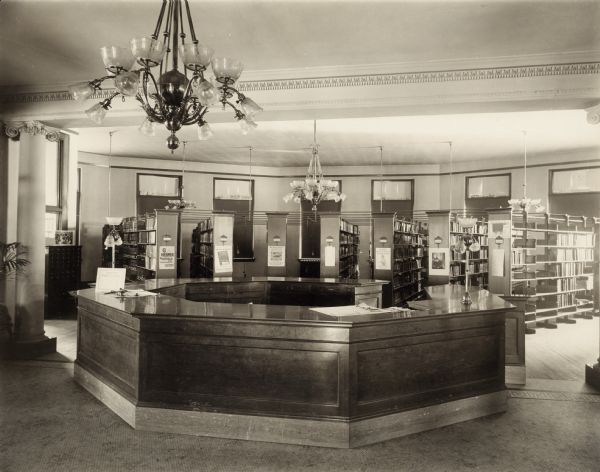 Interior view of the Superior Public Library. Reverse of the cardboard backing reads: "Arch: Carl Wirth, 1902, Carnegie bldg., cost $50,000." In the foreground is a large chandelier. An eight-sided librarian's desk is in the center, framed by columns, beyond which is a room with bookshelves which radiate in a half circle from the desk.