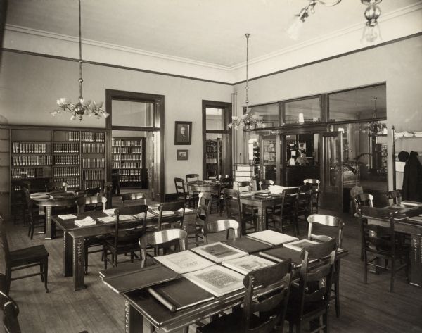 Interior view of the Superior Public Library. One doorway frames a woman (librarian?) working at an eight-sided librarian's desk. A picture of Andrew Carnegie hangs on the wall. In the room are man reading tables with books and other materials displayed on them. Bookshelves line the room. Several doorways lead to other rooms.