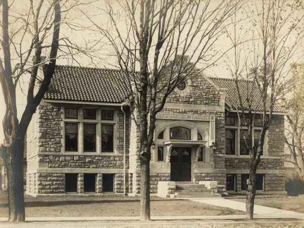 Exterior view of the Waupun Public Library. The library opened in 1905. Reverse of cardboard backing reads: "$12,000." Above the main entrance it reads: "Waupun Public Library," and above that it reads: "Carnegie." Date on corner stone reads: "1904." The stone building has a tile roof, and arched entryway.
