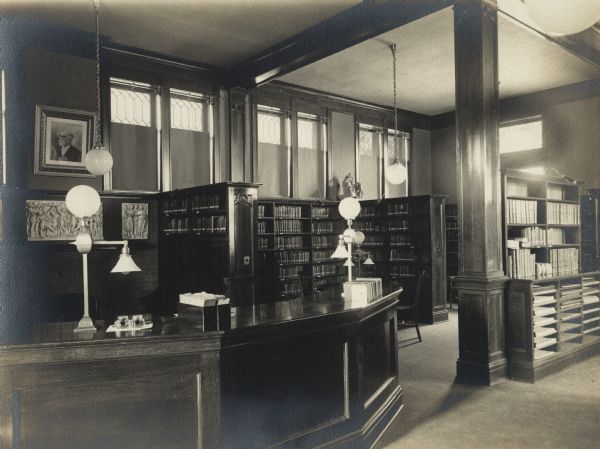 Interior view of the Waupun Public Library. The library opened in 1905. In the foreground is a large librarian's desk, with ink wells, and a card box. Behind the desk is a fireplace  inset with a bas relief. To the right of the desk are bookshelves and tall windows.