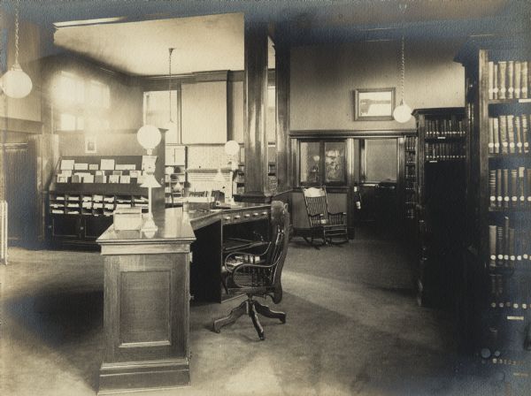 Interior view of the Waupun Public Library. The library opened in 1905. The side of a large librarian's desk is in the foreground. There are bookshelves on the right, a rocking chair near an office in the corner, and a large display rack and reading table on the left.