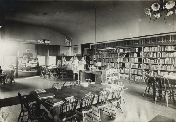 Interior view of the West Bend Public Library. The library was located in the City Hall. The room has an arched ceiling. In the foreground are reading tables with chairs. Bookshelves are along the wall on the right, a 12-drawer card catalog is near the librarian's desk. There are also newspaper racks, chandeliers, and a display board with U.S. flags.
