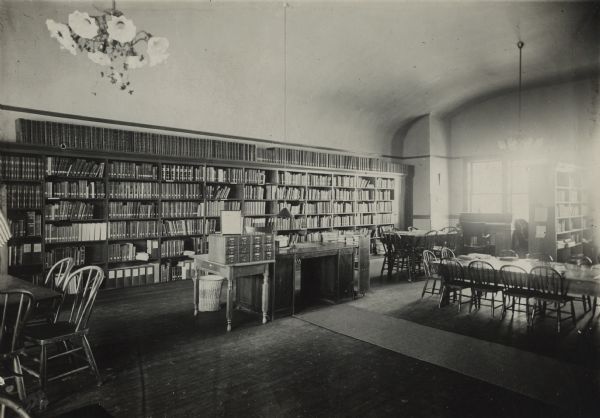 Interior view of the West Bend Public Library. The library was located in the City Hall. Reverse of photograph reads: "Interior West Allis Public Library - In the City Hall." Bookshelves are along the back wall, and there are reading tables and chairs. In the center is a 12-drawer card catalog near the librarian's desk.