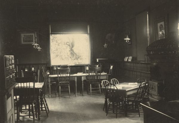 Interior view of the Whitehall Public Library. In the foreground a reading tables and chairs, and a small children's reading table. Bookshelves are along the wall on the right behind a large freestanding cast iron stove, and on the left is a nine-drawer card catalog. A large statue of an angel is set on the sill of a large window.