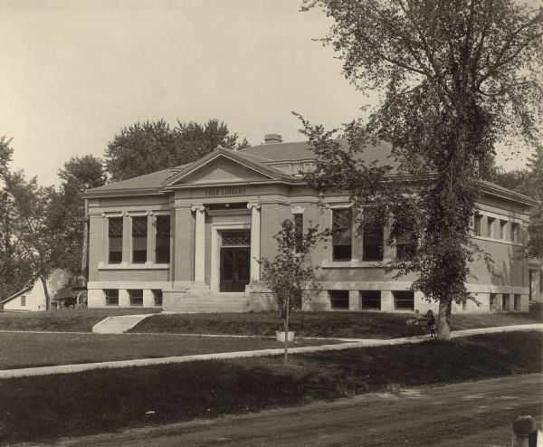 Exterior view from road of the White Memorial Library. The library opened in 1904 and was funded with a $15,000 donation from Flavia White. Above the main entrance it reads: "White Memorial," and above that it reads: "Free Library." The brick building has an entrance with a pediment and two columns. On the sidewalk on the right is a small girl with a toddler in a baby carriage.