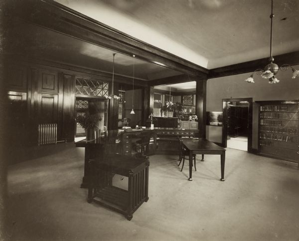 Interior view of the White Memorial Library. The library opened in 1904 and was funded with a $15,000 donation from Flavia White. In the foreground is the back of a large three-sided librarian's desk which faces the entrance, which has wood paneling, and transom windows. There are card catalog drawers in the desk, and bookshelves are on the right wall. Several doorways frame views of other rooms.