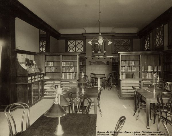 Interior view of the White Memorial Library. The library opened in 1904 and was funded with a $15,000 donation from Flavia White. Bottom right of photograph reads: "Reading & reference room - White Memorial Library, Whitewater, Wis., Claude and Starck Arch'ts." In the room are reading tables with reading lamps and bentwood chairs. Two bookshelves, and a display rack for magazines, divide the room from a reading table in front of a brick fireplace on the back wall which is flanked by bookshelves with windows above.