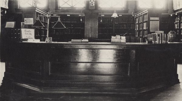 Interior view of the White Memorial Library. The library opened in 1904 and was funded with a $15,000 donation from Flavia White. A large librarian's desk has a stack of pamphlets with a sign placed on them that reads: "Federation Cook Book, 50 cents." Behind the desk is a card catalog, and bookshelves on the back wall below a row of windows.