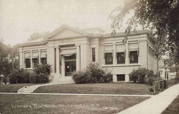 Exterior view of the White Memorial Library. The library opened in 1904 and was funded with a $15,000 donation from Flavia White. Bottom of photograph reads: "Library Building, Whitewater, Wis., 41523." Above the main entrance it reads: "White Memorial," and above that it reads: "Free Library." The entrance has a pediment and columns. The windows have striped fabric awnings.