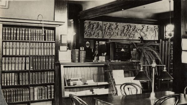 Interior view of the White Memorial Library. The library opened in 1904 and was funded with a $15,000 donation from Flavia White. In the room are reading tables, a newspaper rack, and bookshelves, On the back wall beyond an archway is a bas relief of horsemen. 
