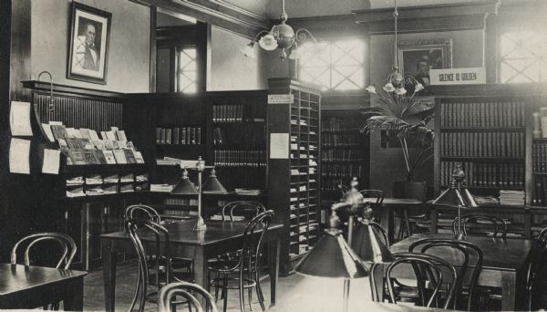 Interior view of the White Memorial Library. The library opened in 1904 and was funded with a $15,000 donation from Flavia White. A sign on a bookshelf reads: "Silence is Golden." In the room are reading tables, bookshelves, chandeliers, and a potted plant. A built-in shelf on the left displays magazines. On the back wall is a brick fireplace flanked by windows and bookshelves.