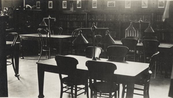 Interior view of the White Memorial Library in Whitewater. The library opened in 1904 and was funded with a $15,000 donation from Flavia White. In the room are reading tables with reading lamps. Bookshelves are along the back wall, and prints are displayed above them.