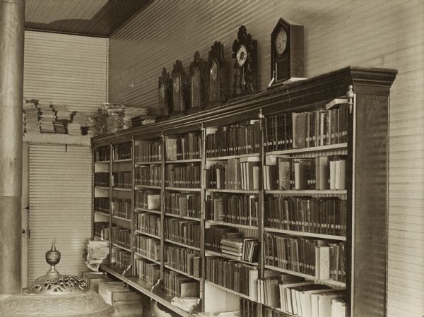 Interior view of the Hillsboro Public Library. Reverse of cardboard backing reads: "Hillsboro Public Library in a Jeweler's store. Take Oct 2, 1905." Bookshelves line the corner of a wall on the right, and six clocks sit along the top. The top of a cast iron stove is in the foreground.