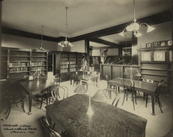 Interior view of the White Memorial Library in Whitewater. The library opened in 1904 and was funded with a $15,000 donation from Flavia White. Bottom left of photo reads: "#1, Whitewater Library, Claude & Starck Archts., Madison, Wis." In the room are reading tables with bentwood chairs, chandeliers, bookshelves, and a 12-drawer card catalog. Native American photographs are along the top of shelves on the right. Other rooms can be seen beyond a doorway and low dividing wall.