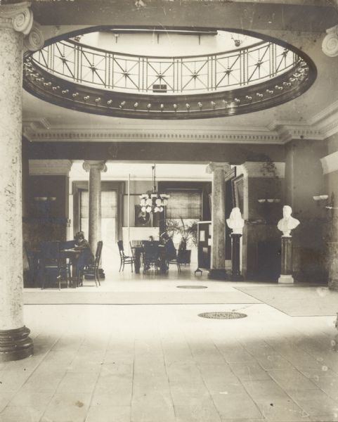 Interior view of the Oshkosh Public Library. On the back is the note: Wm. Walker, Oshkosh, arch. 1900 -- cost $55,000. Ionic marble columns, and a round opening in the ceiling with a railing and perhaps a skylight are in a large open area, and in the background are people sitting at reading tables.