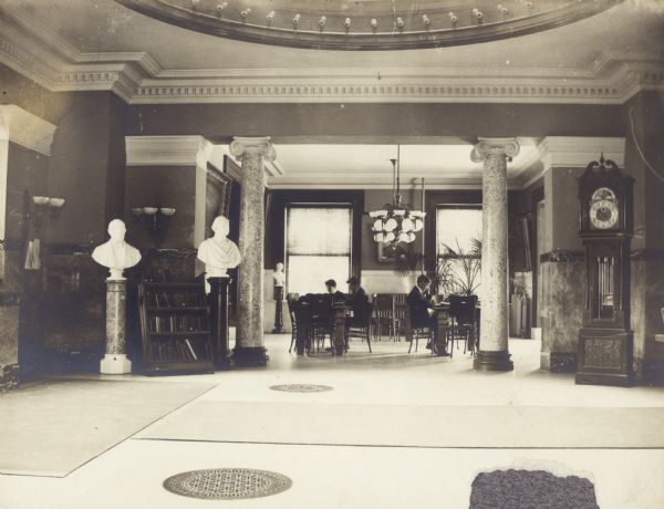 Interior of the Oshkosh Public Library. View is from large open area with Ionic marble columns, with a round opening in the ceiling and perhaps a skylight. In the background are people sitting at reading tables. On the right is a large grandfather clock.