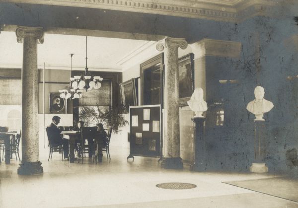 Interior of Oshkosh Public Library. On the back is the note: Wm. Walker, Oshkosh, arch. 1900 -- cost $55,000. View from large open area towards two ionic columns beyond which three people are sitting at a reading table. There is a sculpture on the right, and chandeliers hanging from the high ceilings.