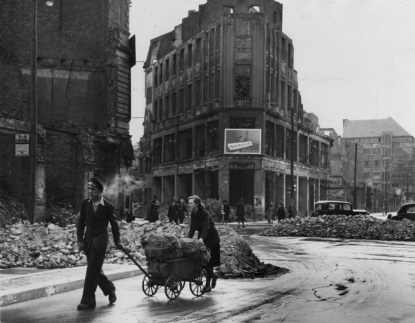 Several days before the Berlin Blockade was to be lifted, Russian authorities permitted this West Berlin couple to purchase potatoes in the eastern sector. Bombed buildings line the street, and piles of bricks and debris litter the sidewalks and spill into the street. The press service caption fails to indicate the reason for the exception. The blockade formally ended on May 11.